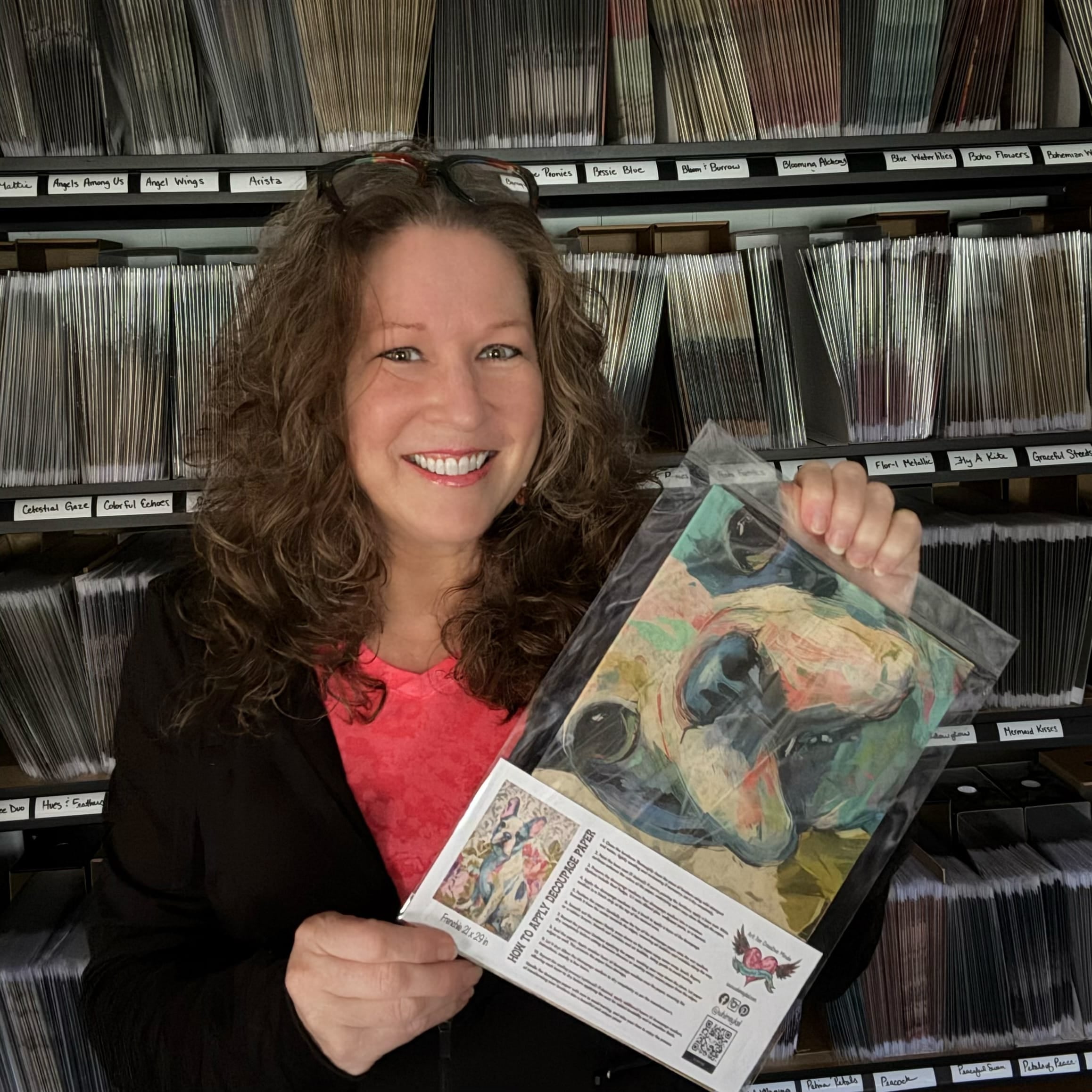 woman holds decoupage paper in a clear bag standing in front of shelves with lots of paper on them