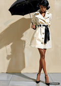 A woman in a cream trench coat with a black belt holds a black umbrella, her shadow cast on the wall.